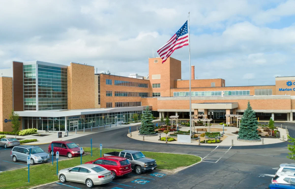 Exterior view of Marion General Hospital campus with a tall American flag, brick buildings, and a circular driveway with parked cars.
