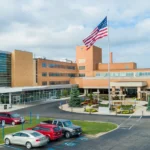 Exterior view of Marion General Hospital campus with a tall American flag, brick buildings, and a circular driveway with parked cars.